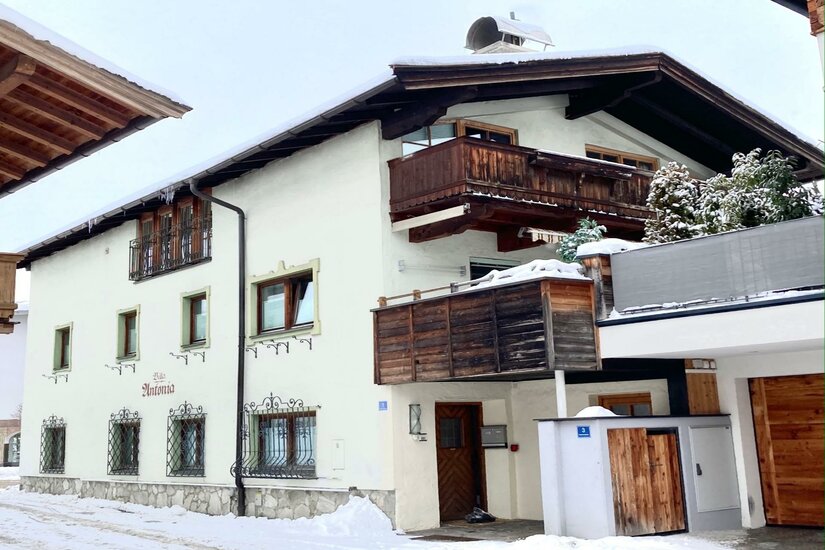 Zweistöckiges weißes Alpenhaus mit Holzbalkonen und schneebedecktem Dach im Winter.