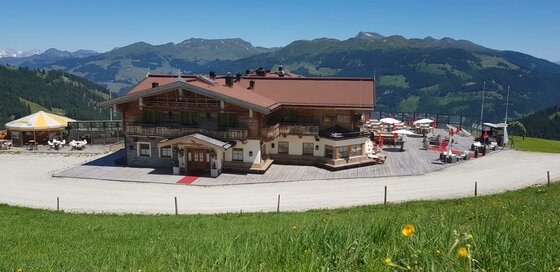 Große Berghütte aus Holz und Stein mit Sonnenterrasse vor einem Gebirgspanorama.