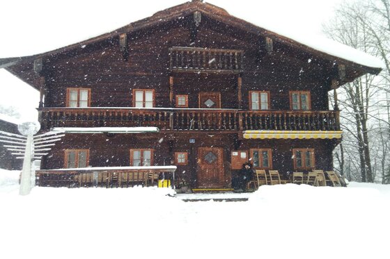 Vorderansicht eines zweistöckigen Holzhauses im Schneefall mit Balkon und Terrasse.