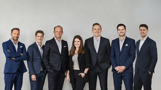 Team Cum Laude Immobilia Sieben Personen in Business-Kleidung stehen nebeneinander vor einem grauen Hintergrund.