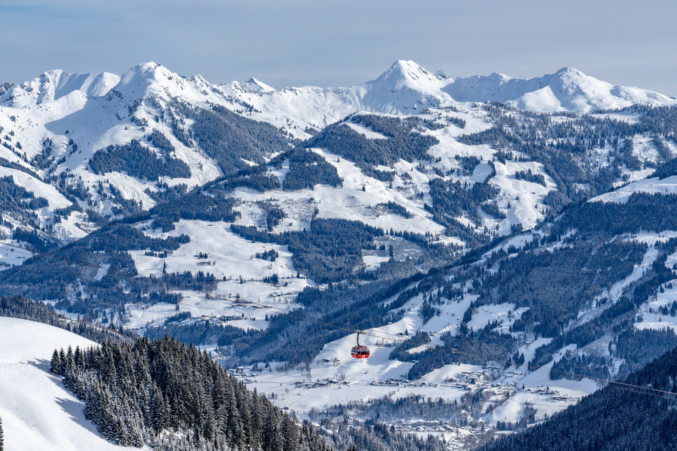 Eine rote Gondel schwebt über schneebedeckte Berge, umgeben von dichten Nadelwäldern und sanften Hügeln.