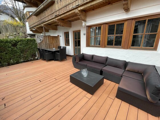 Holzterrasse mit dunklen Loungemöbeln und einem Esstisch vor einem Gebäude im alpinen Stil.