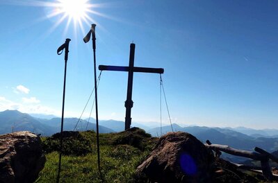 Gipfelkreuz und Wanderstöcke vor Gebirgspanorama bei Sonnenschein.