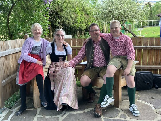Gastgeberfamilie Thainerhof Vierköpfige Familie in traditioneller Tracht sitzt auf einer Holzbank im Freien.