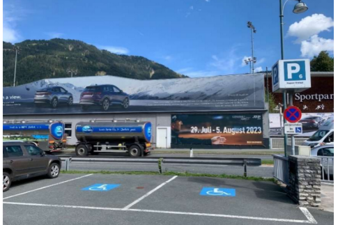 Eine große Werbetafel zeigt zwei Autos vor einer Bergkulisse. Daneben steht ein Parkplatz mit einem Hinweis auf die Parkmöglichkeiten.