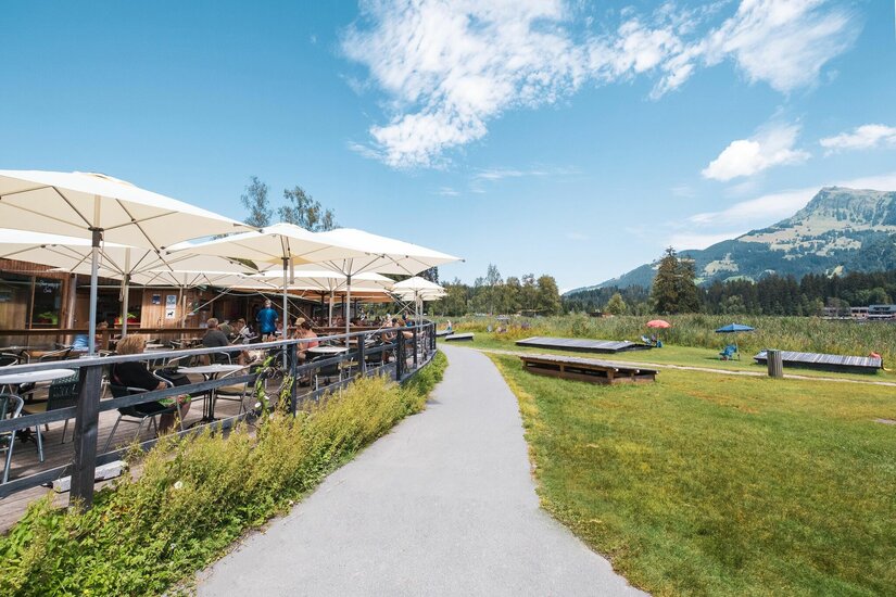 Café Sunshine am Schwarzsee Sonnenterrasse mit weißen Schirmen neben einem asphaltierten Weg und einer Wiese vor Bergkulisse.