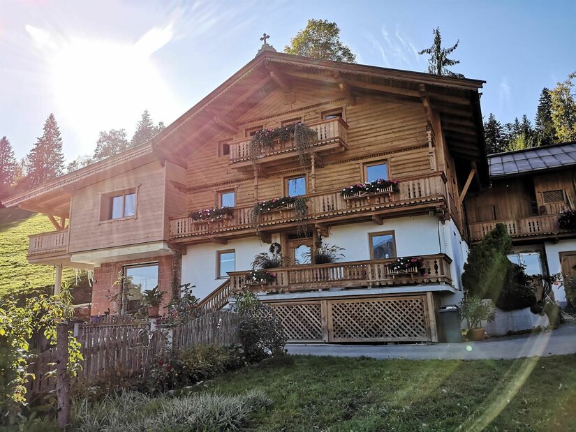 Mehrstöckiges Holzhaus im alpinen Stil mit Blumenbalkonen und weißem Erdgeschoss unter blauem Himmel.