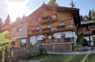 Alpiner Brandhof Mehrstöckiges Holzhaus im alpinen Stil mit Blumenbalkonen und weißem Erdgeschoss unter blauem Himmel.