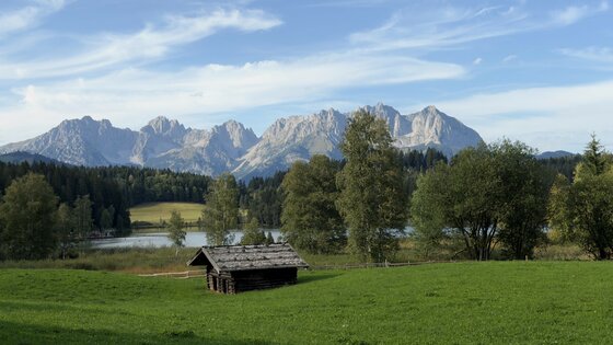Alpenpanorama mit See und Hütte Holzhütte auf einer Wiese vor einem See und einem schroffen Gebirgsmassiv unter blauem Himmel.