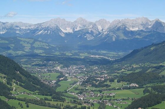 Sommerliches Panorama im Gebirge Blick von einem bewaldeten Hang auf ein Tal mit Siedlungen vor einer markanten, felsigen Gebirgskette.