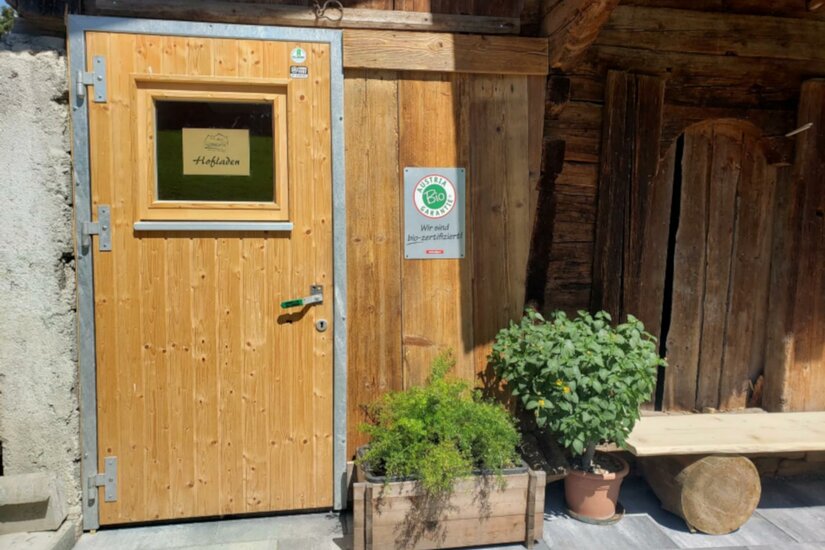 Hofladen Biohof Adler Eine Holztür mit einem kleinen Fenster, umgeben von rustikalen Wänden. Ein Blumentopf mit Kräutern steht vor der Tür.