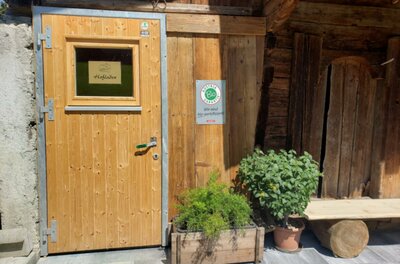 Hofladen Biohof Adler Eine Holztür mit einem kleinen Fenster, umgeben von rustikalen Wänden. Ein Blumentopf mit Kräutern steht vor der Tür.