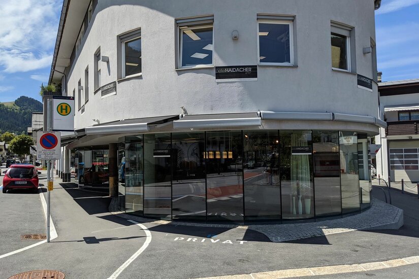 Geschäftsgebäude Peter Haidacher Ein mehrstöckiges Gebäude mit einer großen Glasfront im Erdgeschoss und einer Bushaltestelle davor.