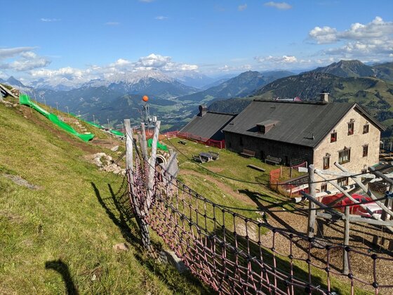 Steinerne Berghütte mit Spielplatz und Rutsche am Grashang vor Gebirgspanorama.