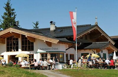 Außenansicht des Hahnenkamm Stüberls mit Gästen auf der Terrasse und Tiroler Fahne unter blauem Himmel.