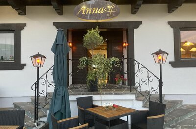 Eingangsbereich des Restaurants Anna mit Außenterrasse, Holztischen, schwarzen Stühlen und beleuchteten Laternen.