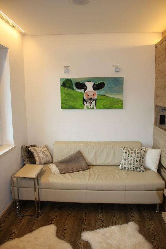 Wohnbereich im Designappartment Jochberg Modernes Wohnzimmer mit hellem Ledersofa, Kuh-Gemälde an der Wand und dunklem Holzboden mit Fellteppichen.