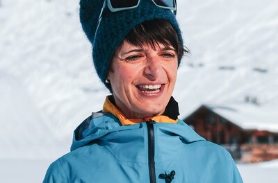 Eine lächelnde Person trägt eine warme Jacke und eine Mütze in den Bergen. Im Hintergrund sind schneebedeckte Hänge zu sehen.