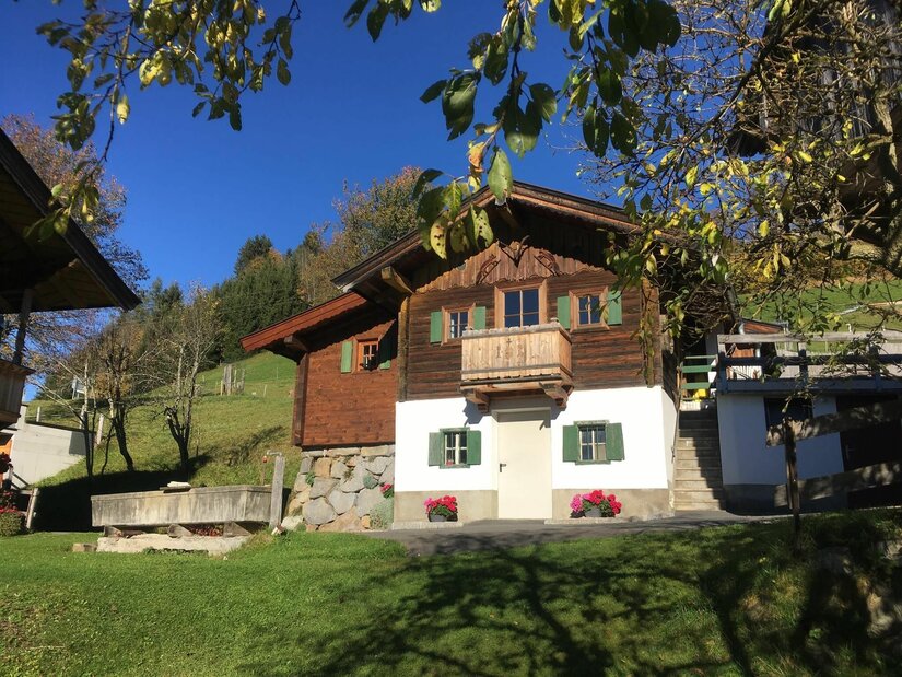 Bauernhof Sinnersberg Hausansicht Alpines Holzhaus mit weißem Sockelgeschoss, grünen Fensterläden und Holzbalkon vor blauem Himmel.