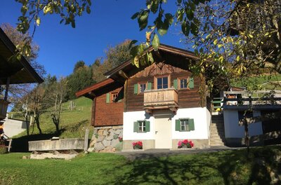 Bauernhof Sinnersberg Hausansicht Alpines Holzhaus mit weißem Sockelgeschoss, grünen Fensterläden und Holzbalkon vor blauem Himmel.