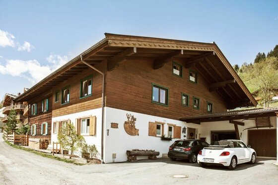 Jagahäusl Jochberg Hausansicht Zweistöckiges Alpenhaus mit dunkler Holzfassade, weißem Erdgeschoss und zwei parkenden Autos davor.