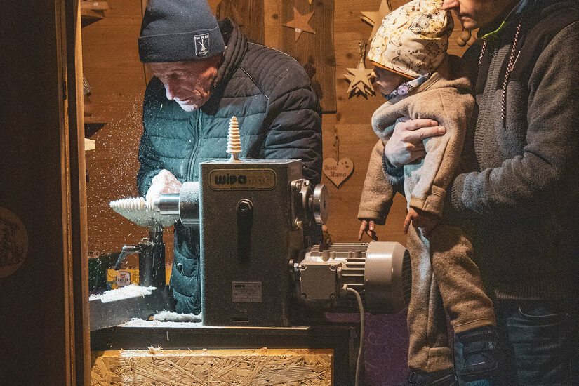 Ein Handwerker schnitzt in einer Holzwerkstatt in Kitzbühel, während eine Familie zuschaut.