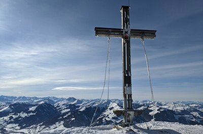 Ein hölzernes Gipfelkreuz auf einem schneebedeckten Berg vor einem Panorama aus verschneiten Alpengipfeln.