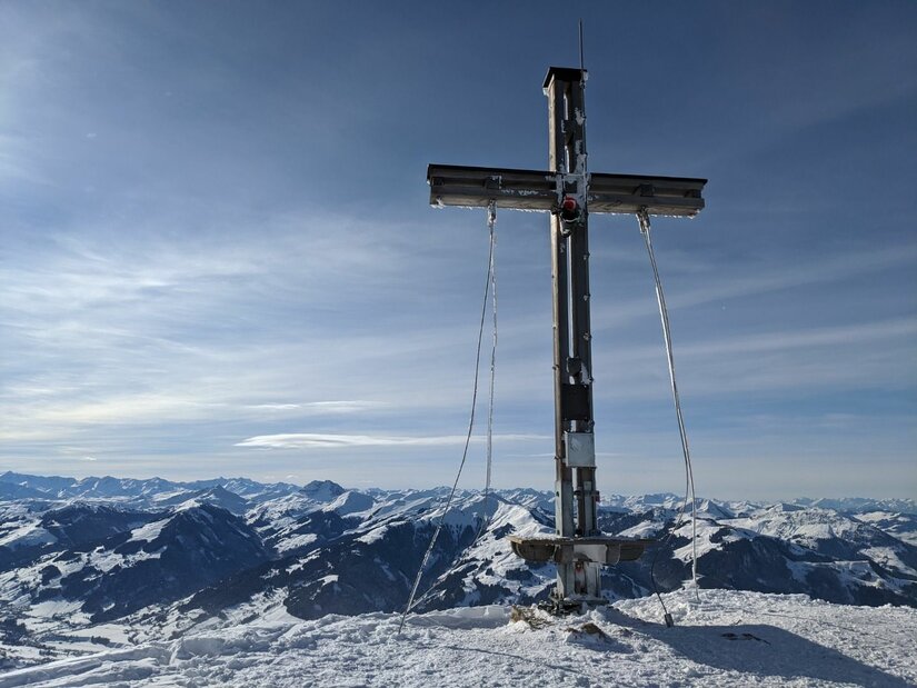 Gipfelkreuz an der Bichlalm Ein hölzernes Gipfelkreuz auf einem schneebedeckten Berg vor einem Panorama aus verschneiten Alpengipfeln.