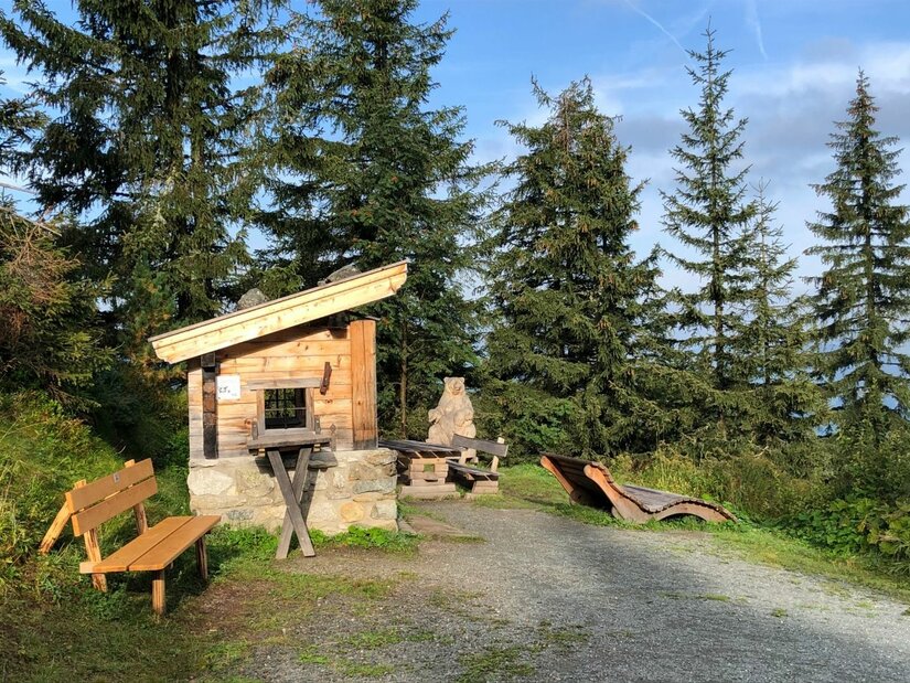 Kleine Holzhütte mit Sitzbänken und einer Holzliege an einem Schotterweg vor Nadelbäumen.
