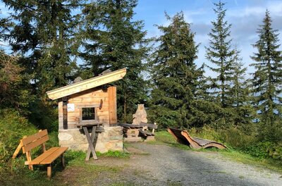 Rastplatz am Streif Spaziergang Kleine Holzhütte mit Sitzbänken und einer Holzliege an einem Schotterweg vor Nadelbäumen.