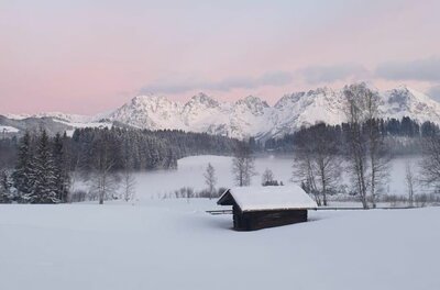 Winterlandschaft am Schwarzsee Verschneite Wiese mit kleiner Holzhütte vor bewaldeten Hügeln und schneebedecktem Gebirgsmassiv unter rosa Himmel.