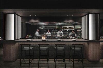 Blick auf eine offene Restaurantküche mit Köchen hinter einem Tresen mit Barhockern.