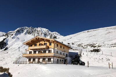Mehrstöckiges Holzchalet in einer verschneiten Gebirgslandschaft unter blauem Himmel.