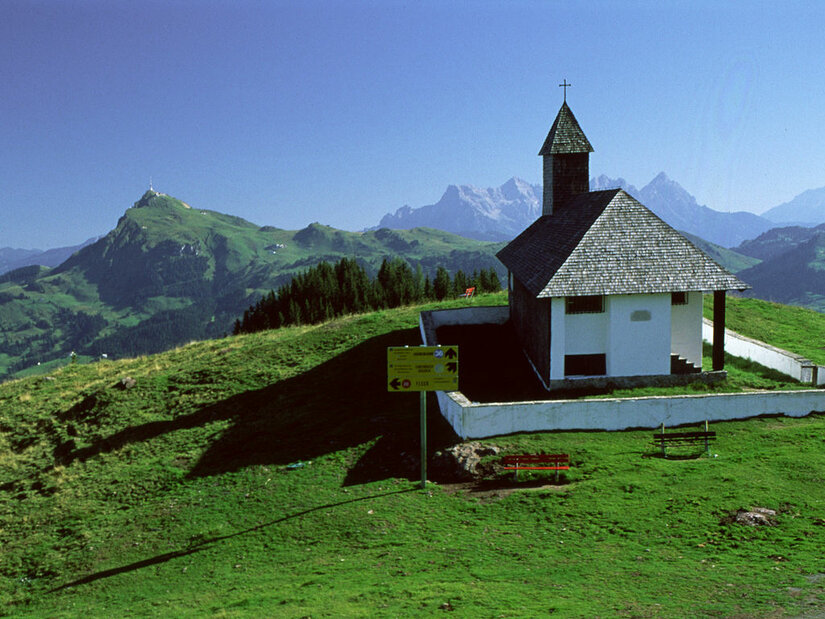 Kleine Kapelle mit Schindeldach auf einem grünen Bergrücken vor alpinem Panorama.