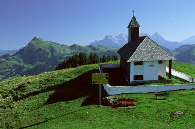 Kleine Kapelle mit Schindeldach auf einem grünen Bergrücken vor alpinem Panorama.