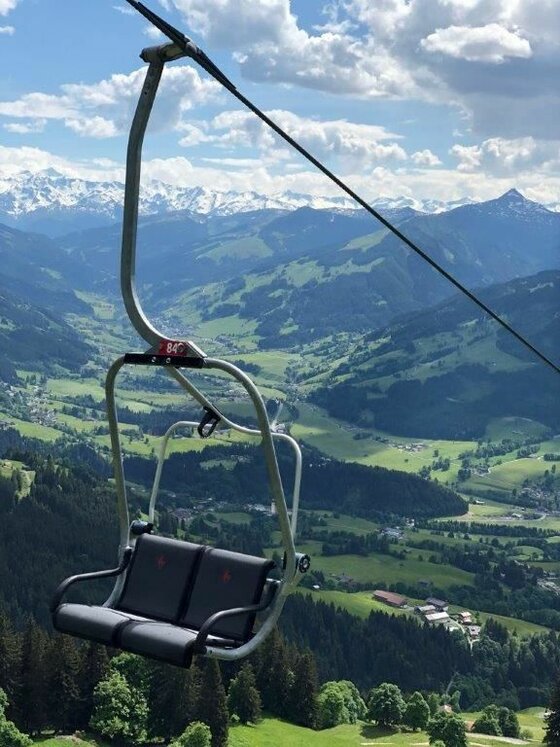 Bichlalmlift in Kitzbühel Ein leerer Zweier-Sessellift schwebt vor einer alpinen Bergkulisse mit grünen Tälern und schneebedeckten Gipfeln.