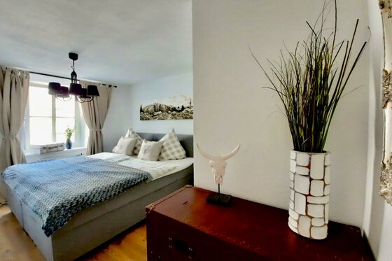 Schlafzimmer mit Doppelbett, Fenster, dunklem Kronleuchter und einer Kommode mit Dekoration im Vordergrund.