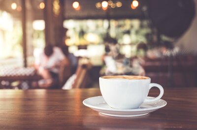Genusscafé <p>Eine Tasse Kaffee auf einem Tisch mit Holzoberfläche. Im Hintergrund sind Personen in einem Café zu sehen.</p>