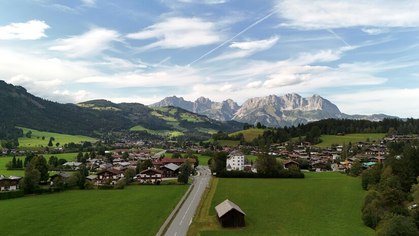 Dorfansicht mit Wildem Kaiser Blick auf ein Dorf im Tal mit grünen Wiesen und dem Gebirgsmassiv des Wilden Kaisers im Hintergrund.