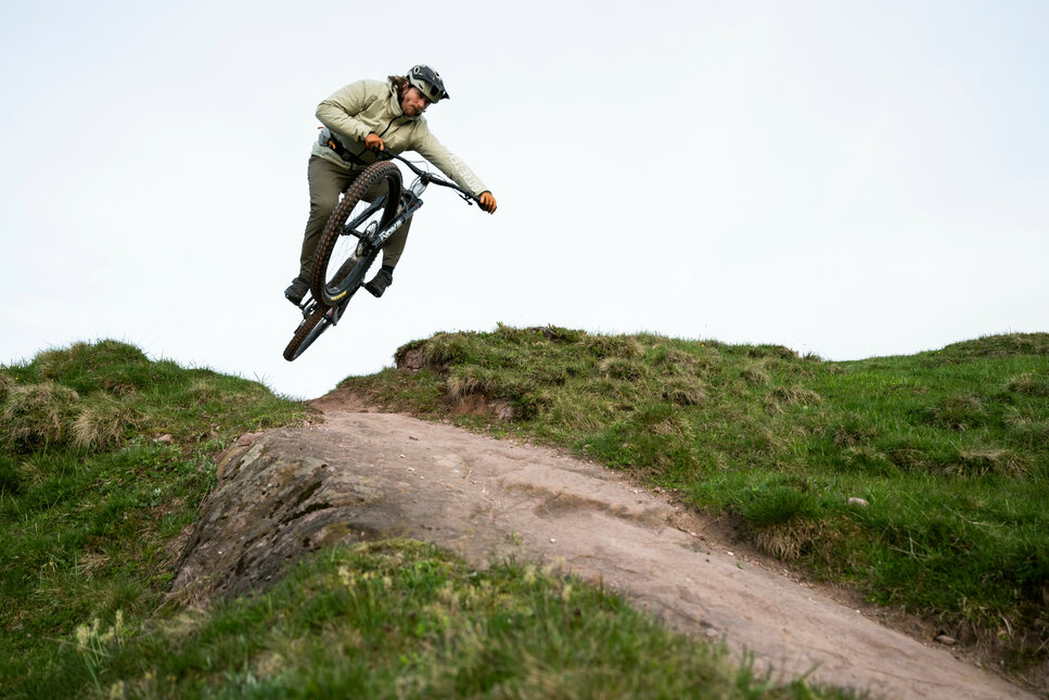 Ein Mountainbiker springt auf dem Hahnenkamm Trail in Kitzbühel über einen Hügel und fährt auf einem schmalen Pfad durch grünes Gras.