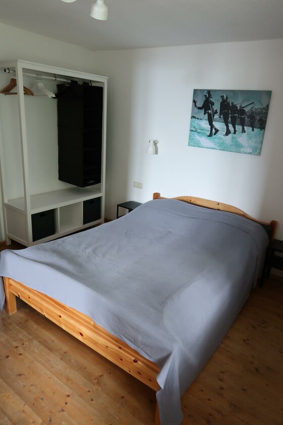 Schlafzimmer mit Holzbett, grauem Überwurf, offenem Kleiderschrank und einem Bild von Skifahrern an der Wand.