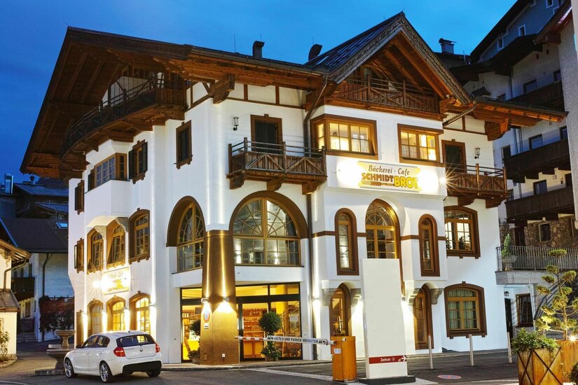 Beleuchtetes Gebäude der Bäckerei Schmidt bei Dämmerung mit weißer Fassade und Holzbalkonen.