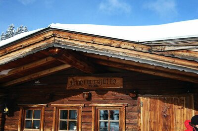 Holzfassade der Jagerwurzhütte mit Namensschild und schneebedecktem Dach unter blauem Himmel.