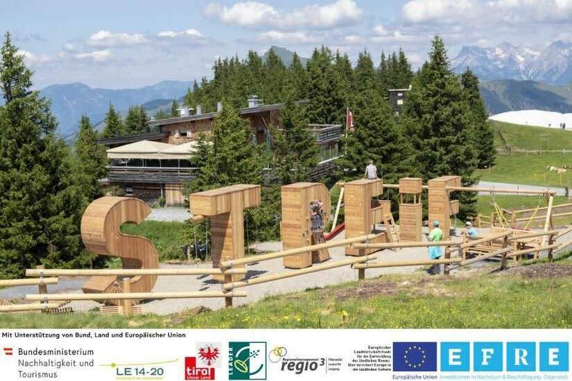 Große Holzbuchstaben als Spielgeräte vor einer Almhütte in den Bergen.
