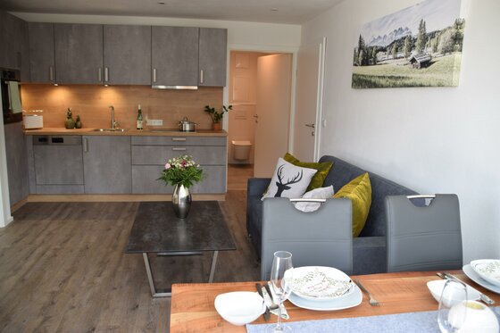 Moderner Wohn- und Essbereich mit grauer Küchenzeile, Sofa und gedecktem Holztisch.
