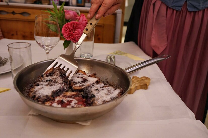 Kaiserschmarrn in der Pfanne Eine Hand serviert mit einem Pfannenwender Kaiserschmarrn aus einer Metallpfanne auf einem gedeckten Tisch.