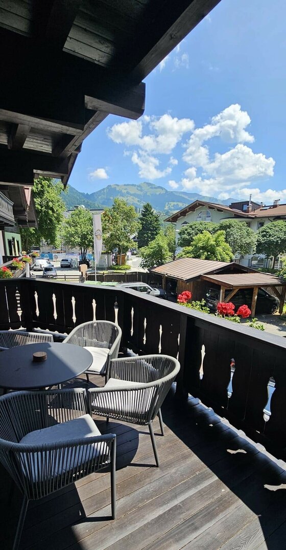 Balkon des Mountain King Chalet Holzbalkon mit modernen Sitzmöbeln und Blick auf Berge und umliegende Häuser unter blauem Himmel.