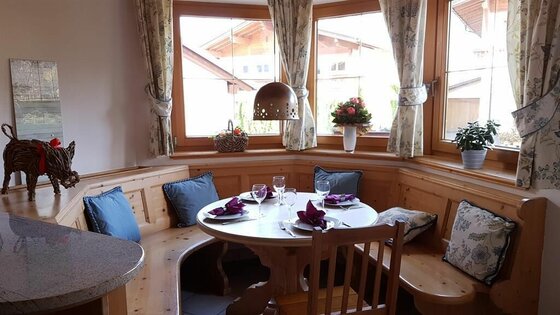 Essecke im Haus Aschenwald Eckbank aus hellem Holz mit rundem Esstisch und blauen Kissen vor einem Fenster.