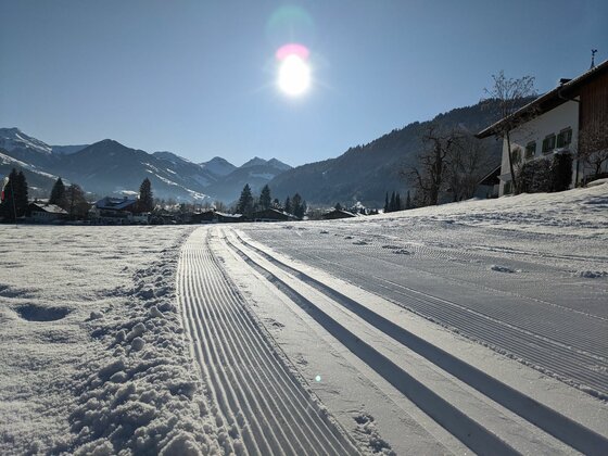 Präparierte Langlaufloipe im Schnee mit Blick auf Berge und Häuser unter sonnigem Himmel.