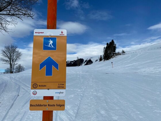 Gelbes Hinweisschild für eine Skitour zur Bichlalm vor einer verschneiten Berglandschaft.